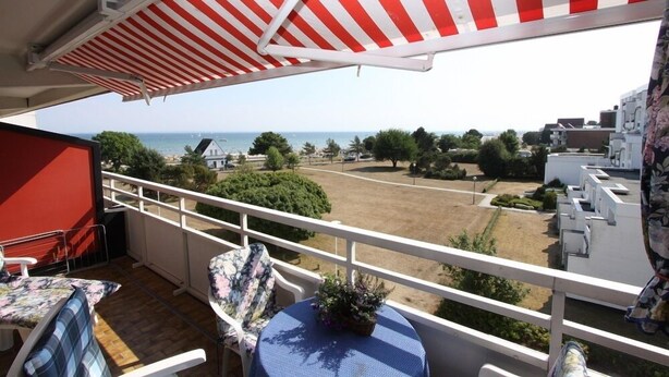 Un balcon agrémenté d’un auvent rayé de rouge et de blanc, une table recouverte d’une nappe bleue, et une vue sur une zone côtière avec des bâtiments et de la végétation.