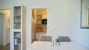 Een eetgedeelte met een witte tafel, grijs gestoffeerde stoelen, een kleine potplant en een glazen vaas met bloemen.