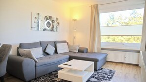 Een moderne woonkamer met een grijze hoekbank, een witte salontafel en een vloerlamp. Er is een groot raam met uitzicht op bomen.