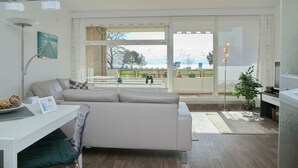 A modern living room with a sofa, a desk, and a large window offering a view of a balcony. - Goldene Ruh App. 103 Inkl. Wlan (Grömitz)