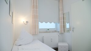 A small, clean room with a single bed, a bedside table, a radiator, a small cabinet, and a window with curtains. - Goldene Ruh App. 103 Inkl. Wlan (Grömitz)