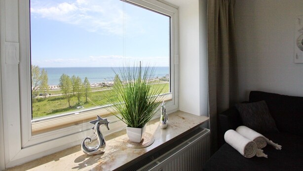 Une pièce dotée d’une grande fenêtre donnant sur une plage, une plante en pot posée sur le rebord de la fenêtre, un radiateur et un canapé de couleur sombre sur lequel sont pliées des couvertures.