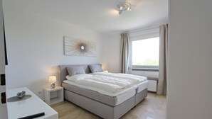 Une chambre à coucher comprenant un lit, des tables de chevet, une lampe, un tableau décoratif et une fenêtre avec des rideaux.
