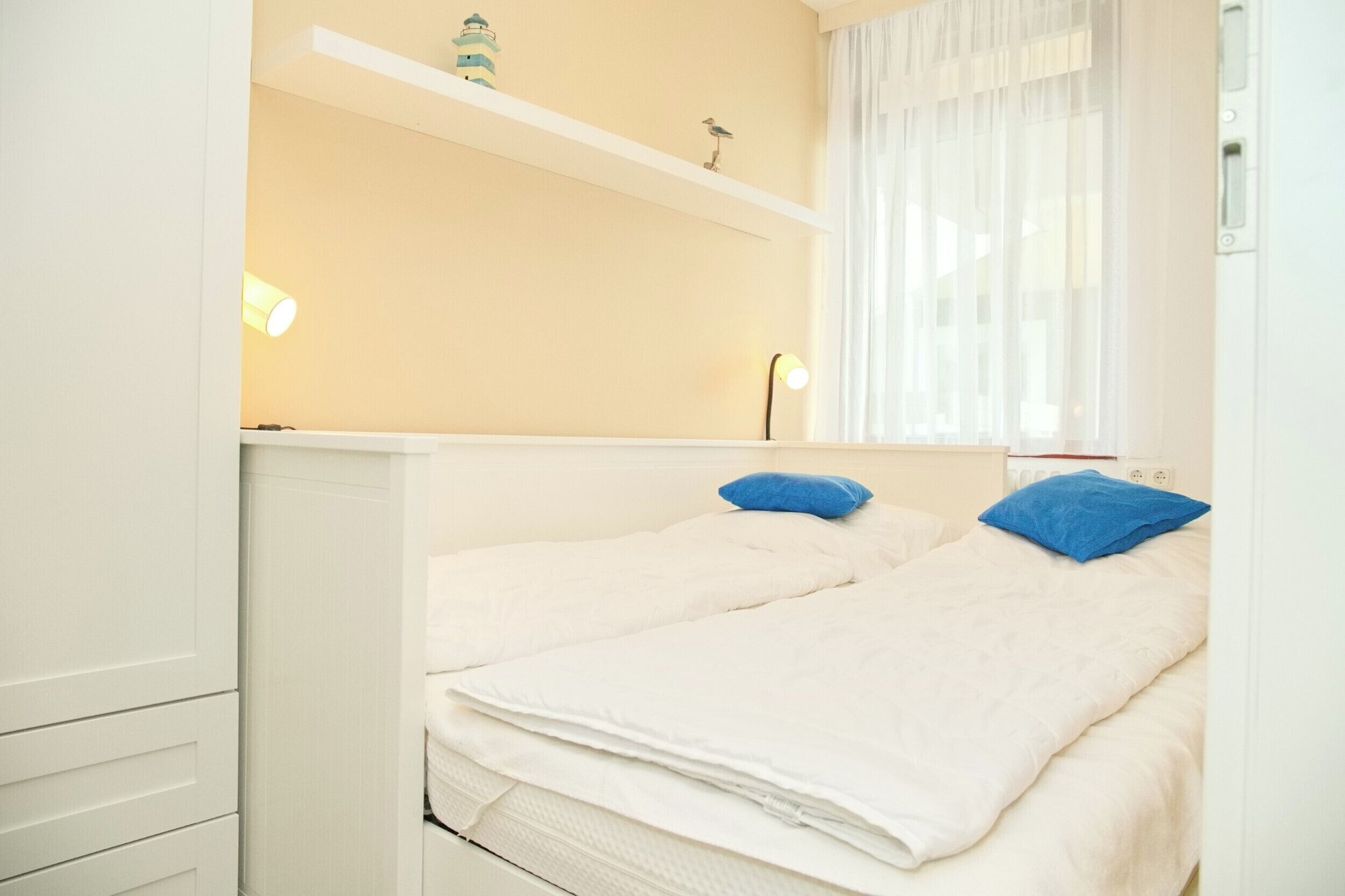 A small, neatly made bed with white bedding and two blue pillows, a shelf with decorative items, and a window with sheer curtains.