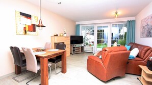 A living room with a wooden dining table, two sofas, a TV, and a large window with curtains. - Möwenstraße Haus 1 Inkl. Wlan (Grömitz)