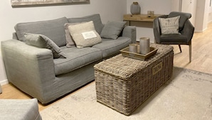 En stue med en grå sofa, en flettet skammel, et lille træ sidebord og en grå lænestol.