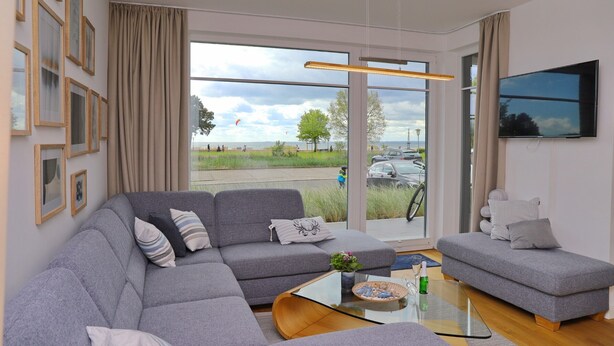 A modern living room with a grey sectional sofa, a small grey loveseat, and a glass coffee table. There is a flat-screen TV mounted on the wall and a large window offering a view of the outdoors. - Sealoft THE ONE Inkl. Wlan (Grömitz)