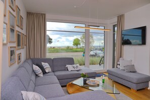 A modern living room with a grey sectional sofa, a small grey loveseat, and a glass coffee table. There is a flat-screen TV mounted on the wall and a large window offering a view of the outdoors.