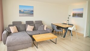 A modern living room with a grey sectional sofa, a wooden coffee table, and a dining table with chairs. There are two wall-mounted pictures, one of a marina and another of a sunset. - Miramar App. 617 Inkl. Wlan (Grömitz)