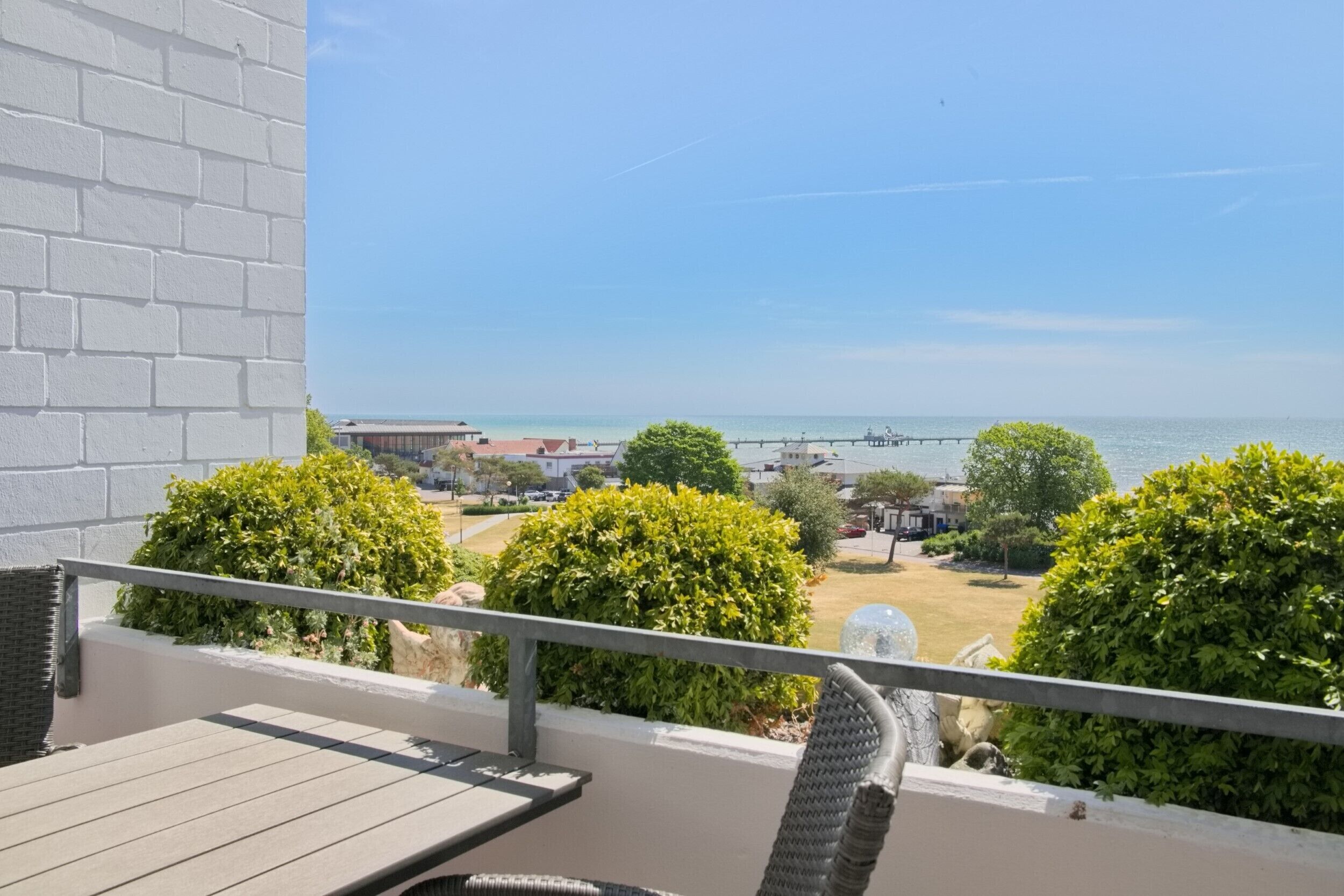 A balcony with a view of the sea, a white brick wall, and green bushes.