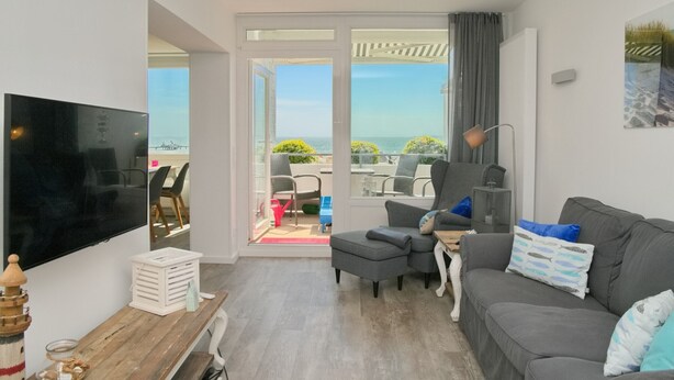 A living room with a grey sofa, a TV, and a view of the sea through large windows. - Tannenburg App. 508 Inkl. Wlan (Grömitz)