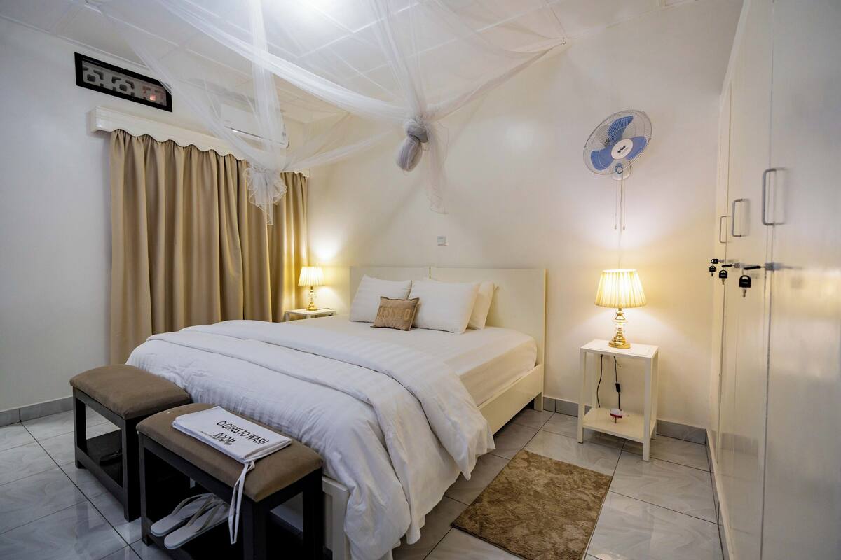 Une chambre d’hôtel comprenant un lit, des tables de chevet, un banc, un ventilateur et une armoire.