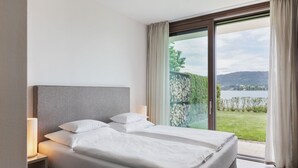 A modern bedroom with a large bed, a nightstand, and a view of a lake through a glass door. - Linde Villa am Wörthersee mit Privatem Strand (Maria Wörth)
