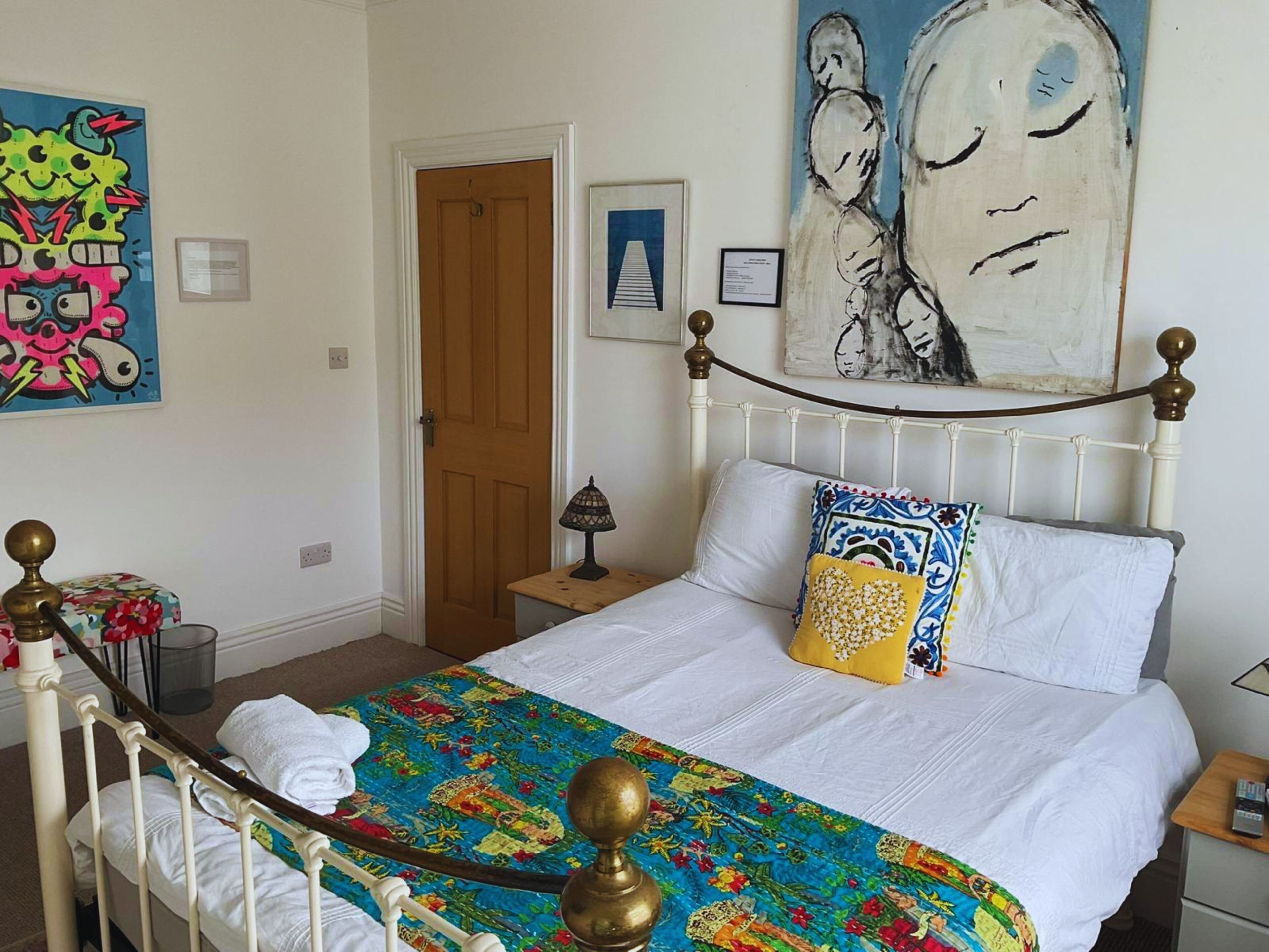A bedroom with a bed, a nightstand, a lamp, a door, and artwork on the walls.