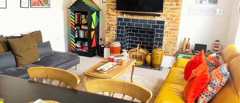 A living room with a sofa, a yellow couch, a TV mounted on a brick wall, a bookshelf, and a fireplace.