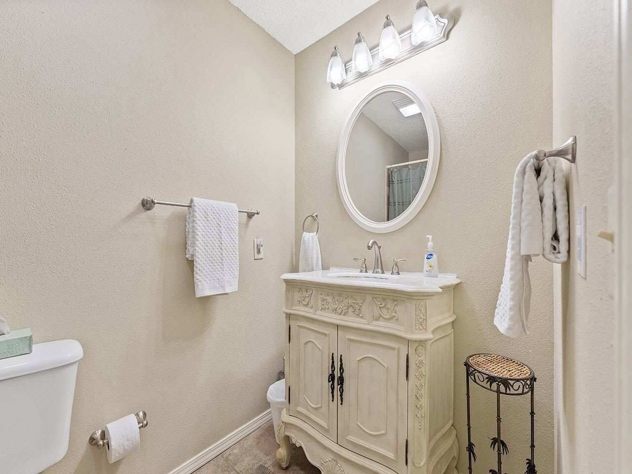 A bathroom with a toilet, sink, vanity, mirror, towel rack, and a small wicker stool.