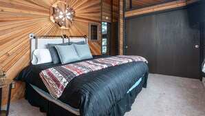 Ein Schlafzimmer mit Holzboden, einem Bett mit gemustertem Überwurf, einem Nachttisch und einer dunklen Tür.