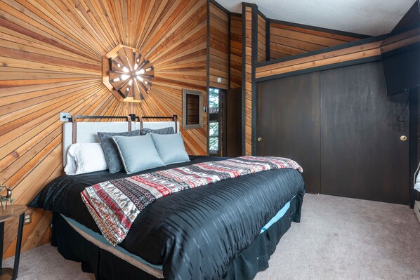 Une chambre avec un plafond en bois, un lit recouvert d’une couvre-lit à motifs, une table de chevet et une porte sombre.