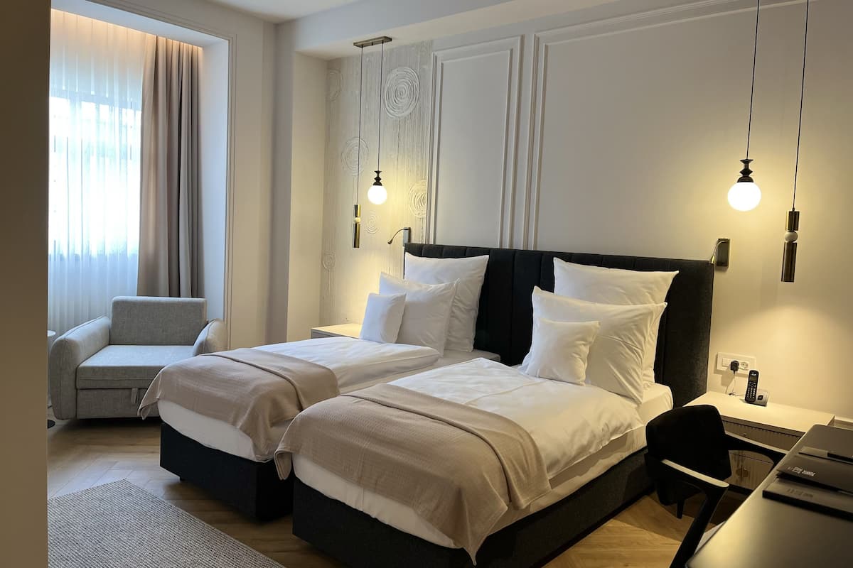 A hotel room with two beds, a desk, a chair, a sofa, and a large window with curtains.