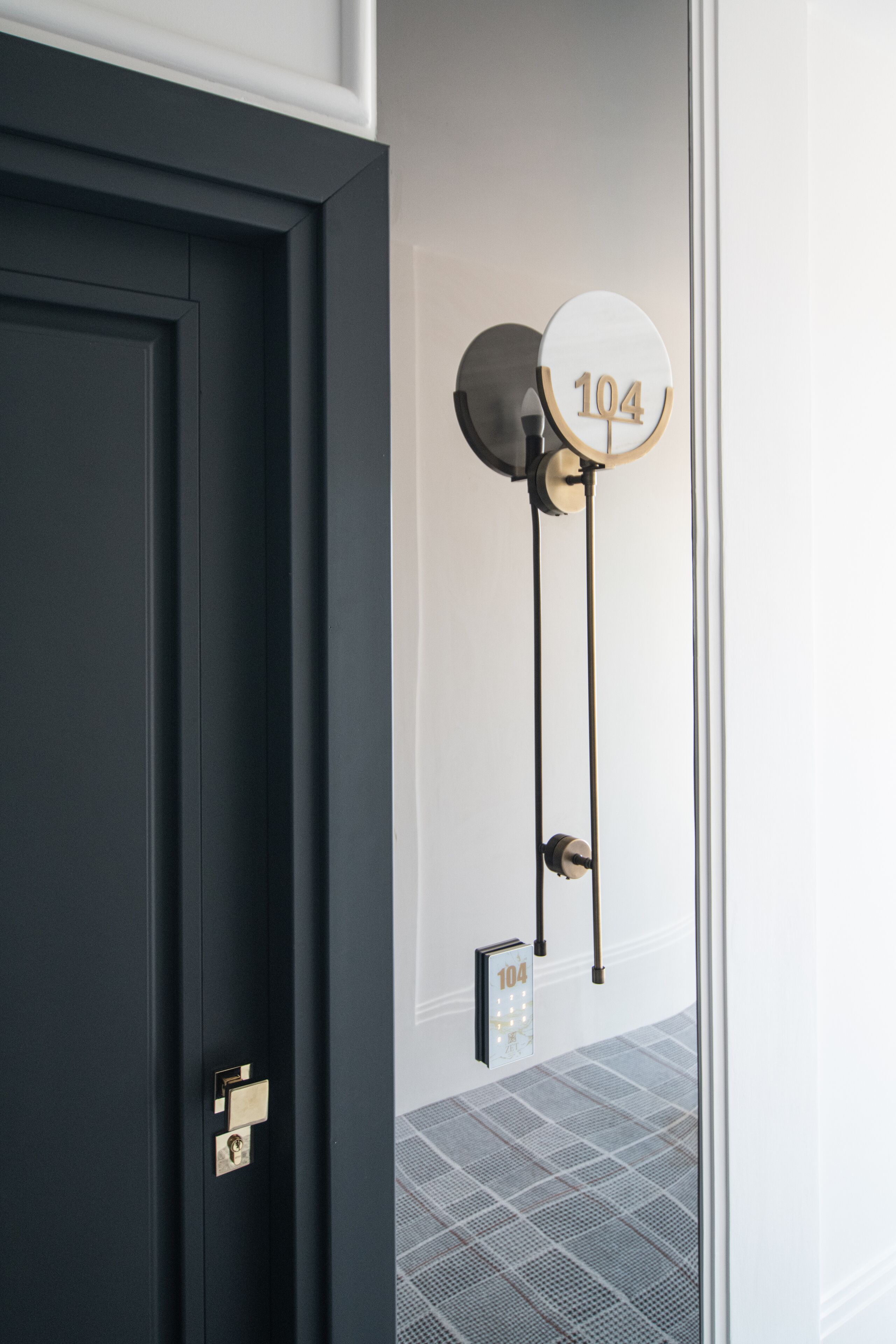 Une porte sombre, dotée d’une serrure en laiton et d’une plaque numérotée « 104 » fixée au mur.