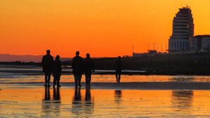 Un capvespre sobre una platja amb gent caminant, un edifici alt al fons i una mar tranquil·la.