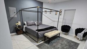 A modern bedroom with a four-poster bed, a bedside table with a lamp, a bench, a ceiling fan, and a window with blinds. - Beautiful apartment 9 min to Mercedes outlets (Weslaco)