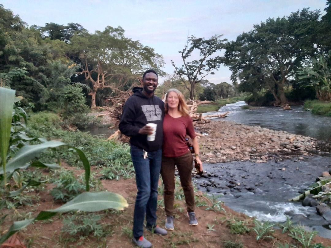 Two people standing by a river with a natural landscape in the background.