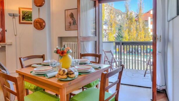 Zona pranzo con tavolo apparecchiato per quattro persone, sedie, un vaso con fiori e vista su alberi e montagne attraverso una porta vetrata.