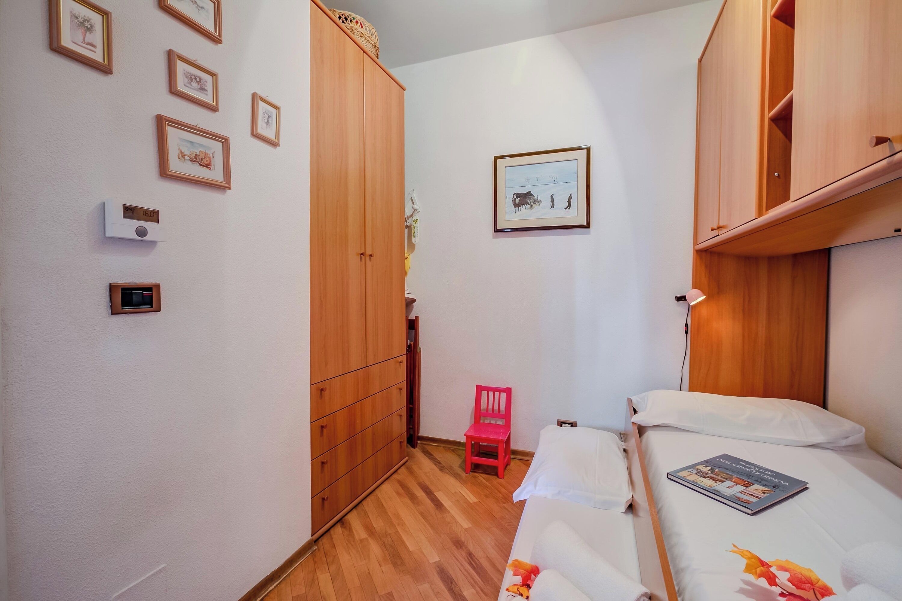 A bedroom with a bed, a red chair, a wardrobe, and a wall-mounted thermostat.