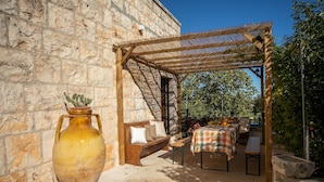 Un patio al aire libre con un banco de madera, una mesa con mantel a cuadros y un jarrón grande con un cactus.