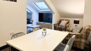 Ein zeitgemäßer Essbereich mit Tisch, Stühlen und einer Topfpflanze. Im Hintergrund befinden sich ein Fernseher und ein Sofa.
