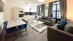 Ein modernes Wohnzimmer mit Sofa, Couchtisch und Essbereich. Im Hintergrund befindet sich eine Küche mit Schränken und Geräten.
