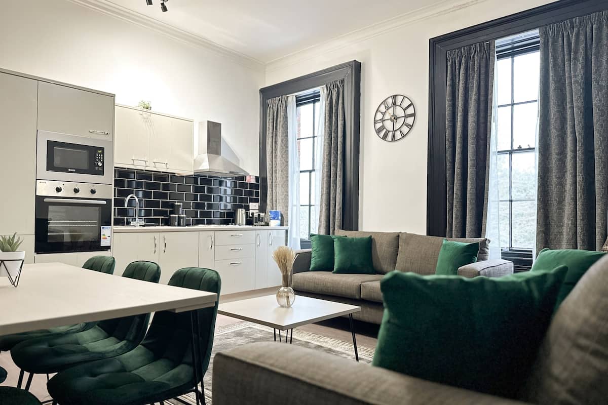 Uma cozinha moderna com eletrodomésticos embutidos, uma área de refeições com mesa e cadeiras, um sofá com almofadas verdes e janelas grandes com cortinas.