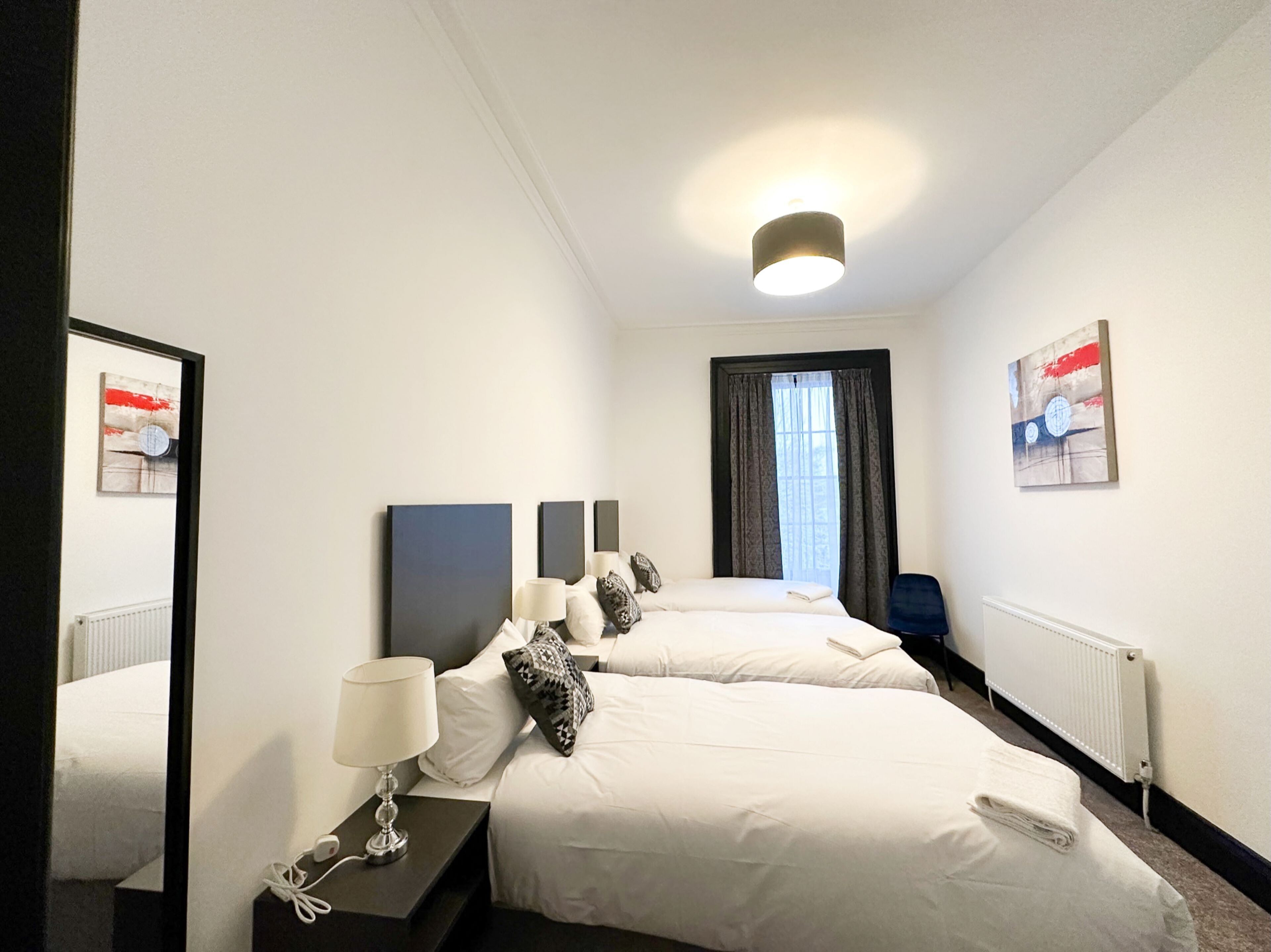 A hotel room with two beds, a nightstand with a lamp, a mirror, and artwork on the walls.