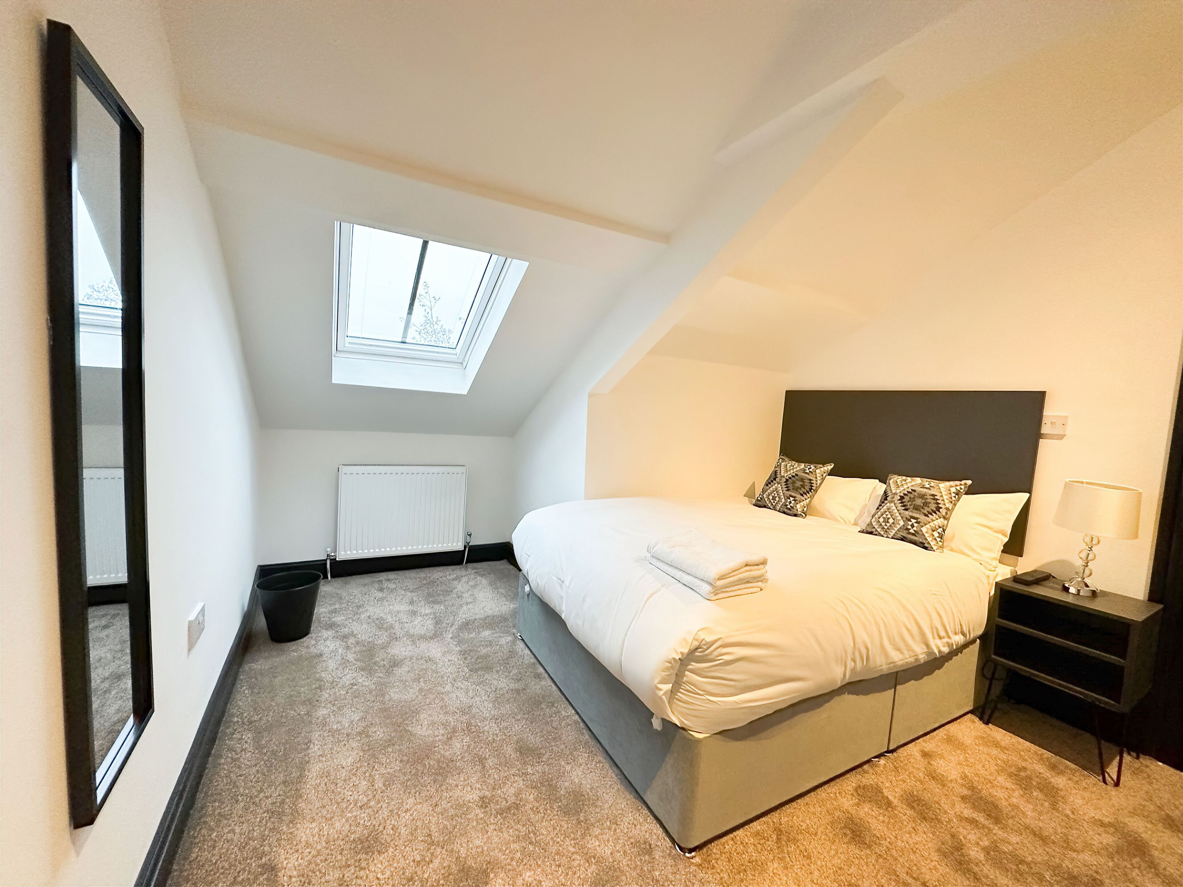 Ein Schlafzimmer mit einem großen Bett, einem Nachttisch, einer Lampe, einem Fenster und einer Heizung.