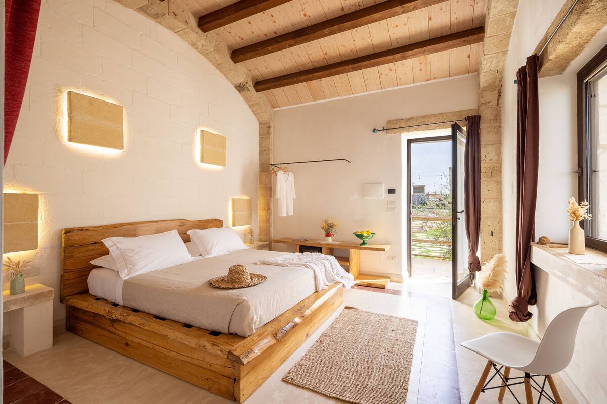 A bedroom with a wooden bed, a white nightstand, a bedside table, a chair, and a view of the outdoors.