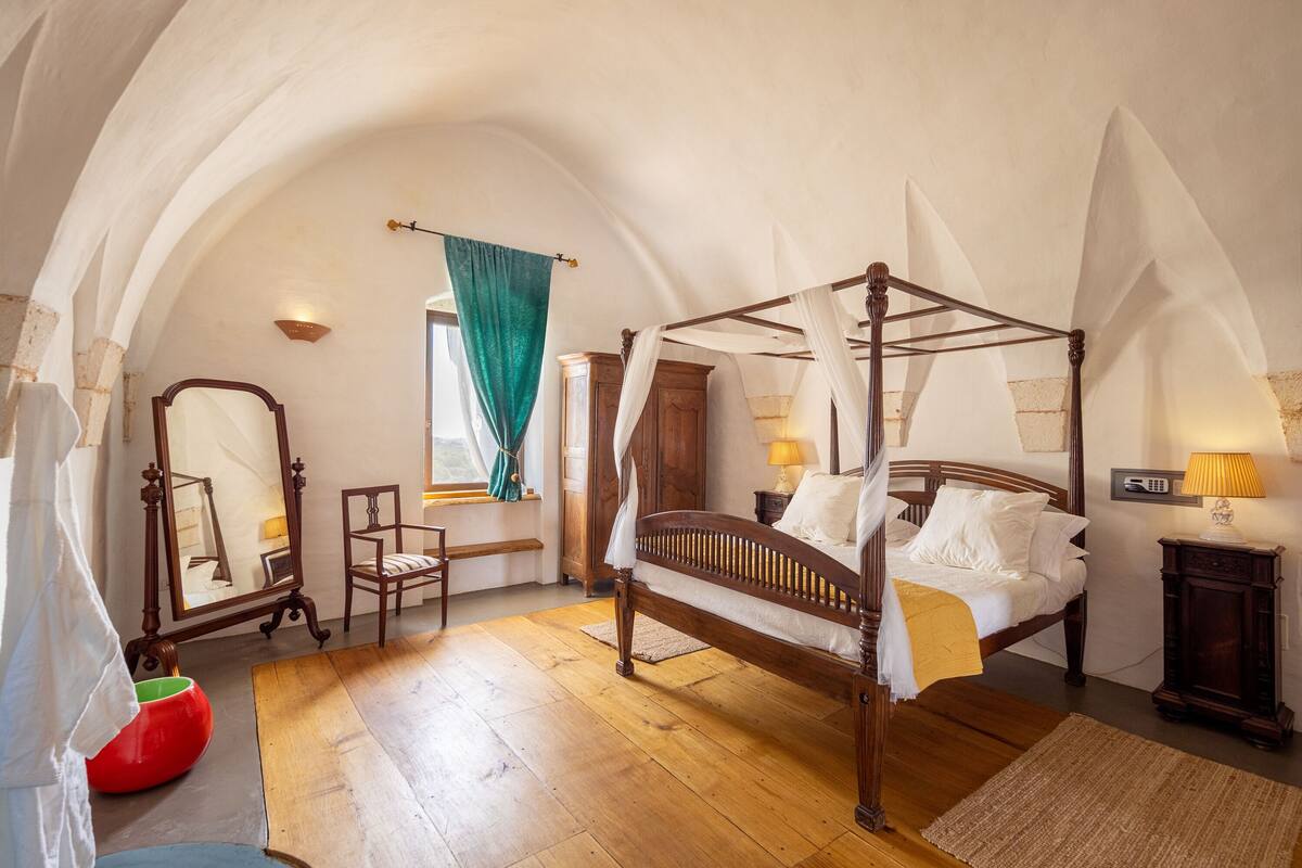 A four-poster canopy bed in a room with wooden floors, a mirror, a chair, and a window with curtains.