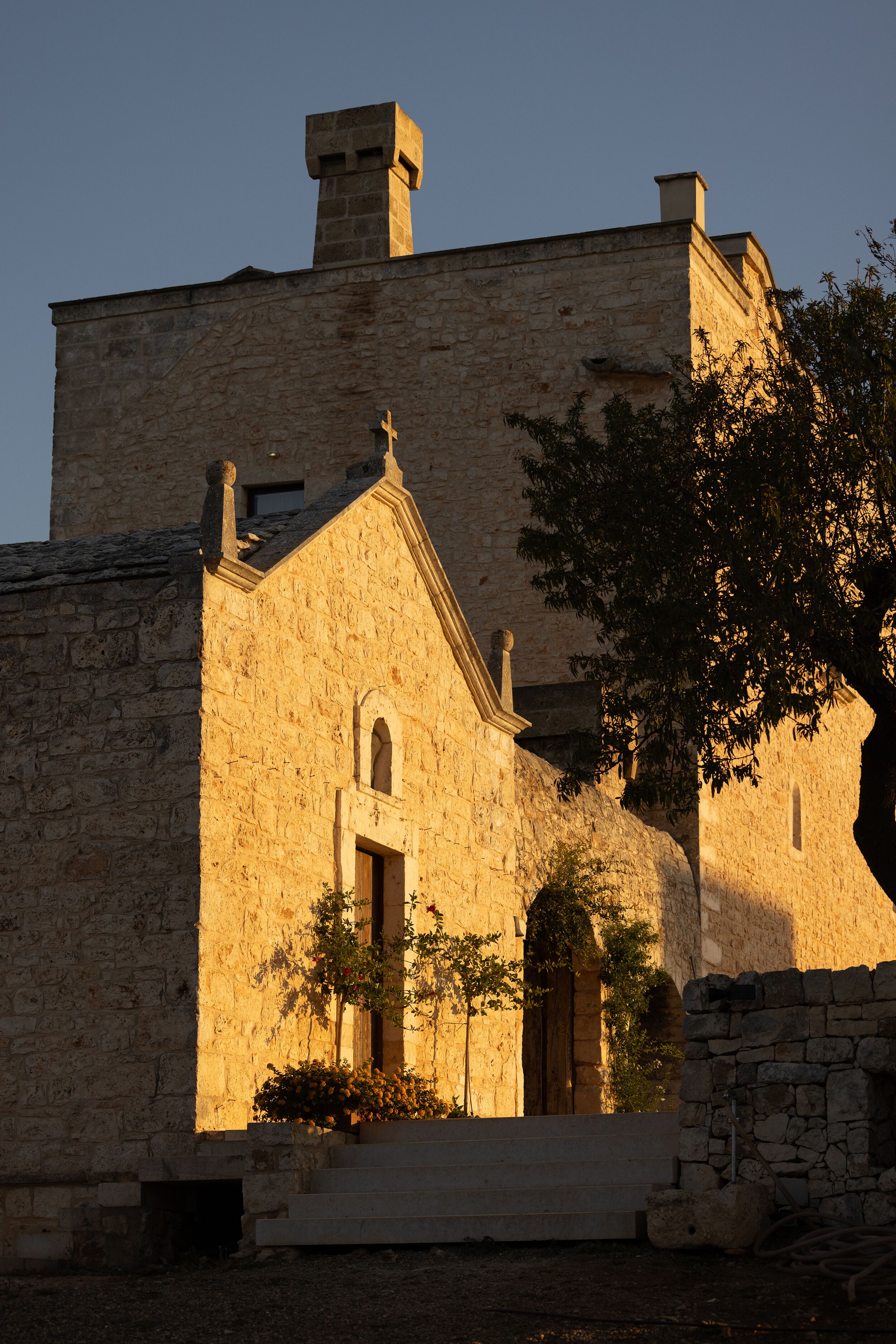 Un edificio de piedra con un arco pronunciado y una ventana pequeña, rodeado por un muro de piedra y un árbol.
