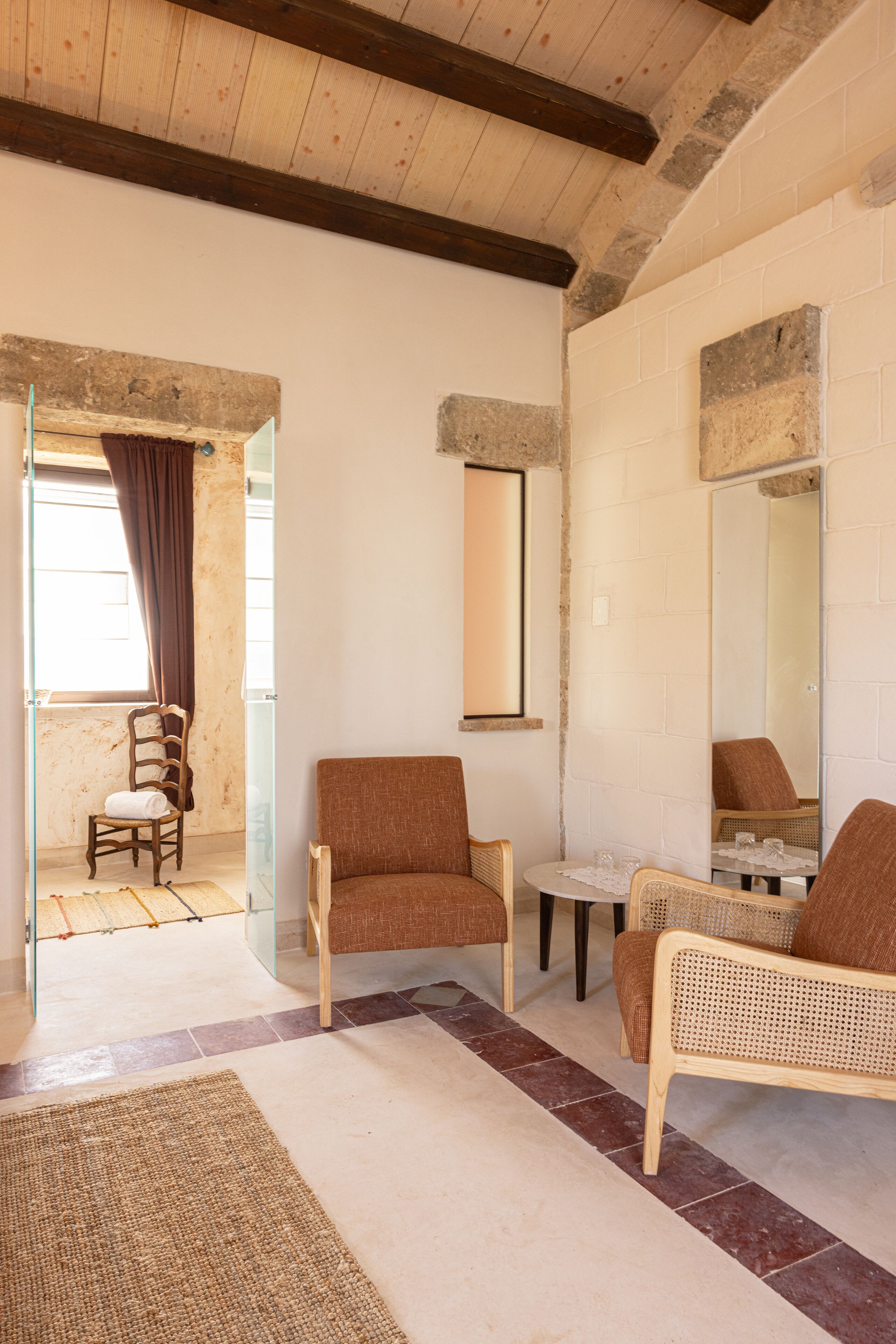 A room with a wooden ceiling, a glass door leading outside, a brown sofa, a wicker chair, and a small table with a mirror.