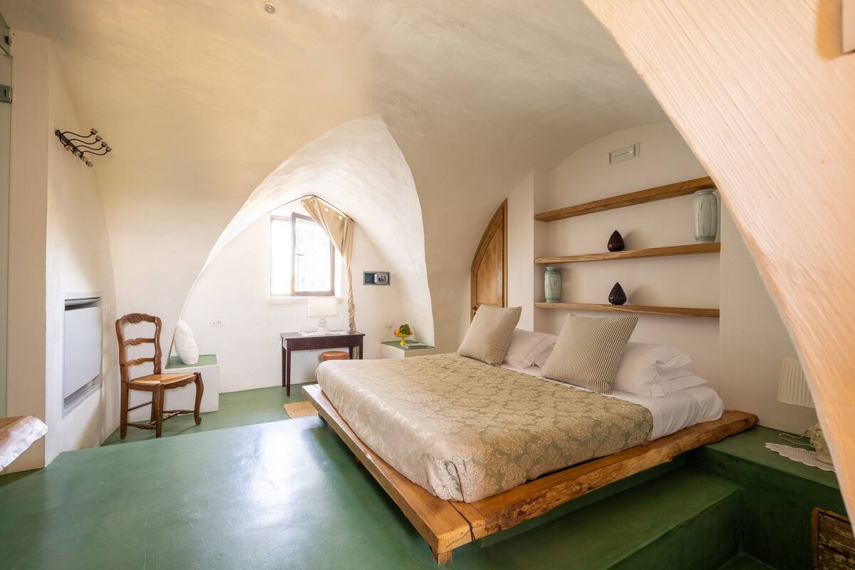A bedroom with a wooden bed, a desk, and a chair. There is a window with curtains, a shelf with decorative items, and a green floor.