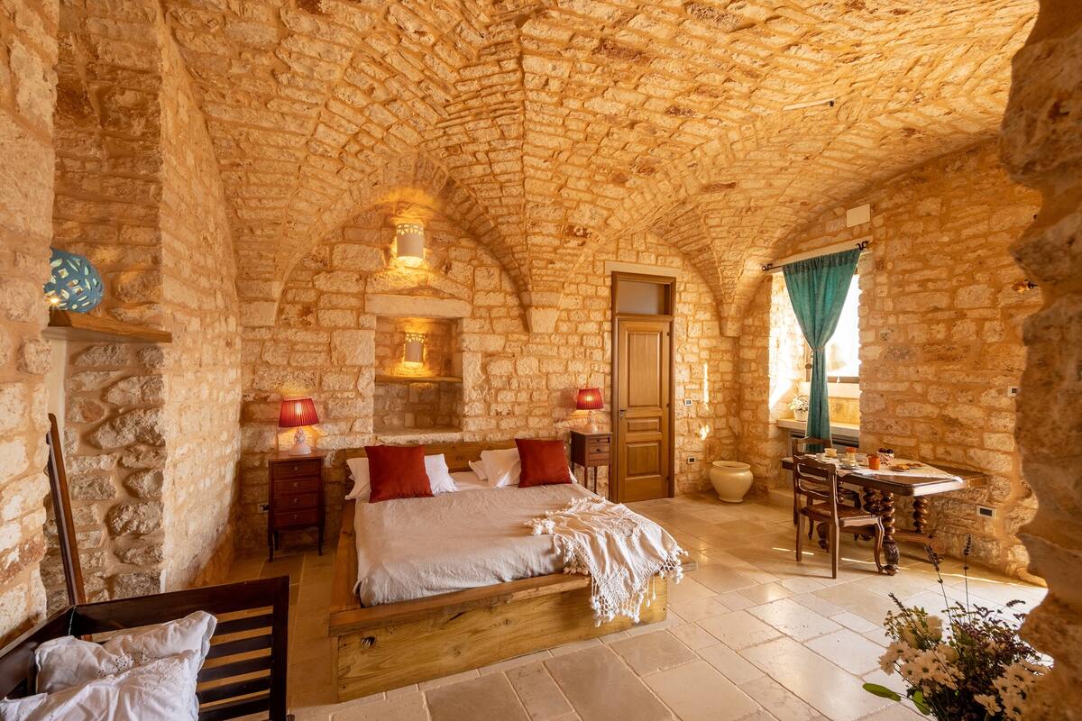 A stone-walled bedroom with a wooden bed, a small table, and a chair.