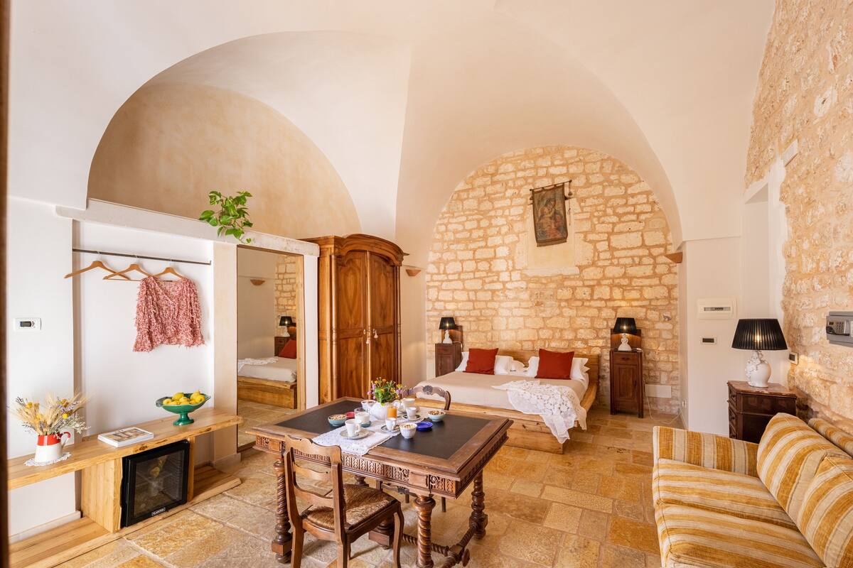 A cozy room with a stone wall, a wooden bed, a dining table with chairs, and a sofa.