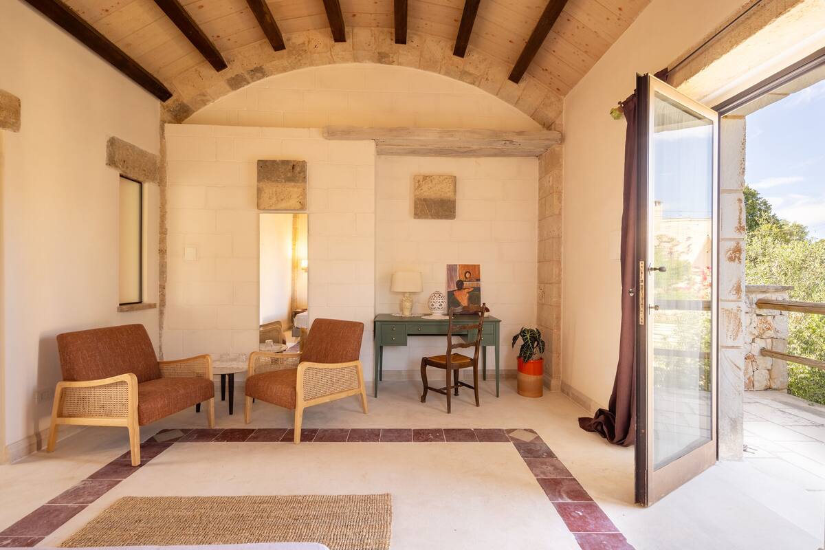 A cozy room with a stone arch ceiling, a wooden desk, two wicker chairs, a small table, and a potted plant.