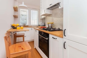 A modern kitchen with white cabinetry, a wooden countertop, and a built-in oven. There is a window with blinds, a hanging light fixture, and a bowl of fruit on the counter.