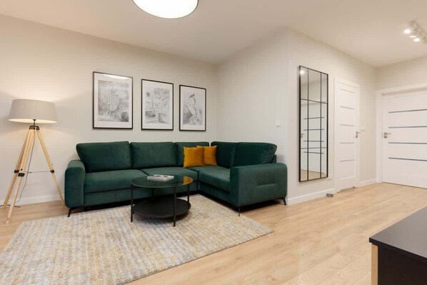 Una sala de estar moderna con un sofá verde, una mesa de centro y una lámpara. Hay tres cuadros enmarcados en la pared y un espejo grande en la puerta.