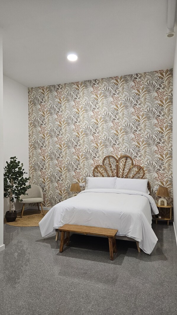 A bedroom with a bed, a bench, a chair, a potted plant, and a patterned wallpaper.