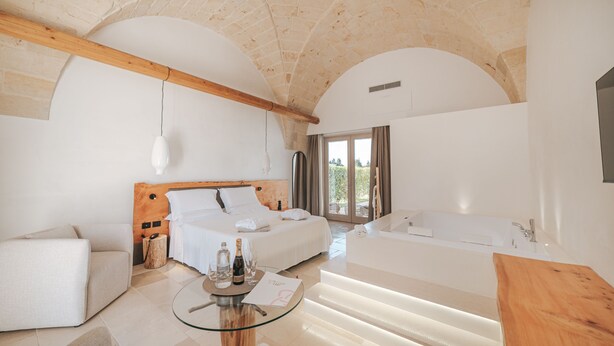 Una habitación de hotel moderna con una cama grande, una zona de estar, una mesa de vidrio con una botella y vasos, y un banco de madera.