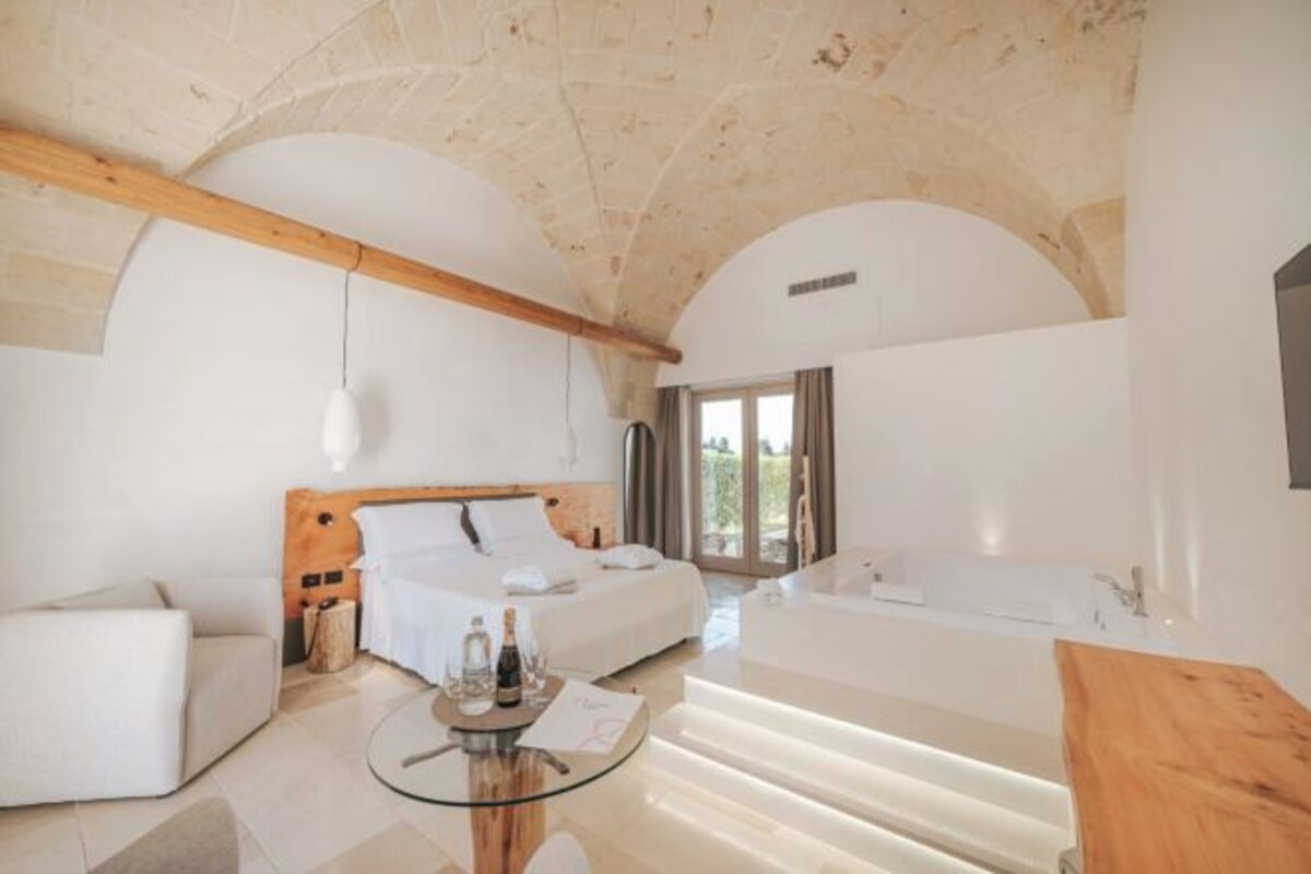 Una habitación de hotel moderna con una cama grande, una zona de estar y una mesa de vidrio con una botella y vasos.
