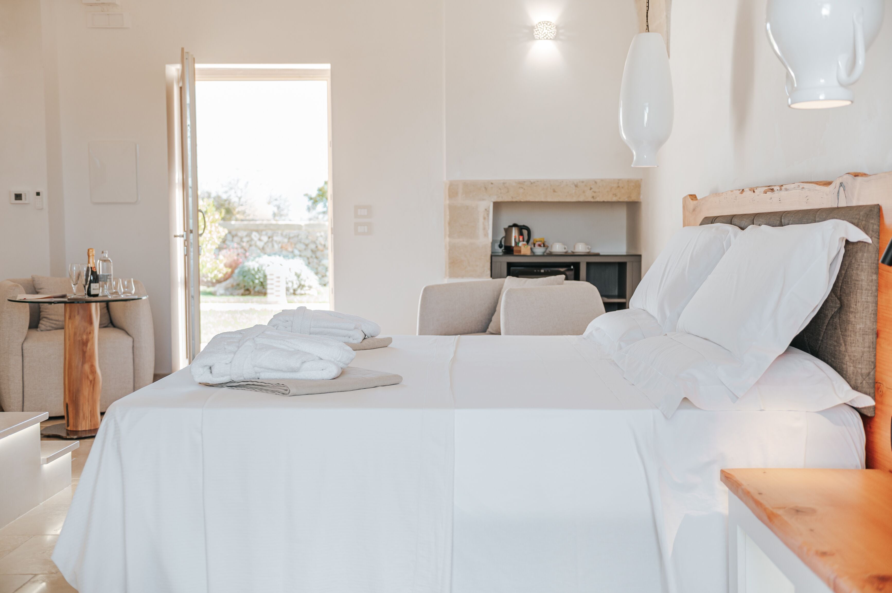 A hotel room with a bed, a small table with a bottle and glasses, a sofa, and a kitchen area in the background.