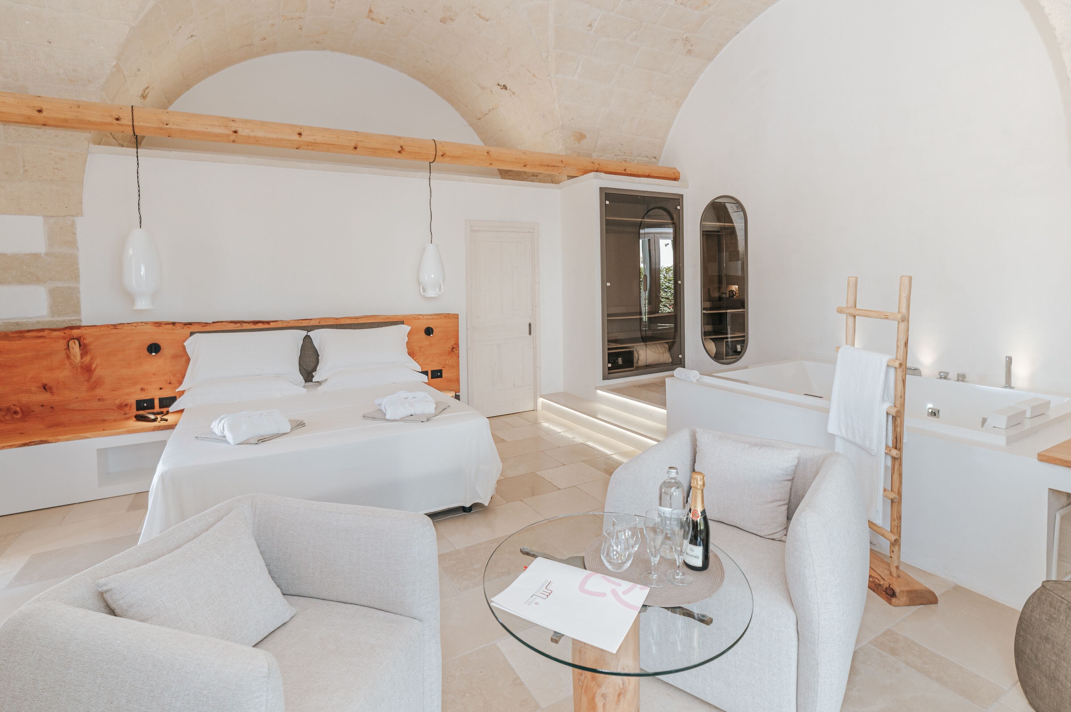 A modern hotel room with a wooden headboard bed, a sitting area with a glass table, and a bathroom with a bathtub and a wooden ladder.